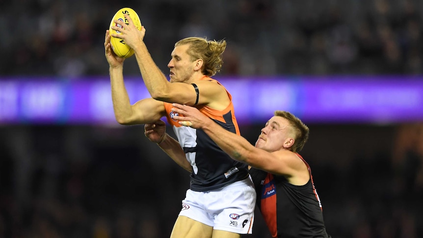 An AFL defender grasps the football above his head while an opponent tries to grab him from behind.