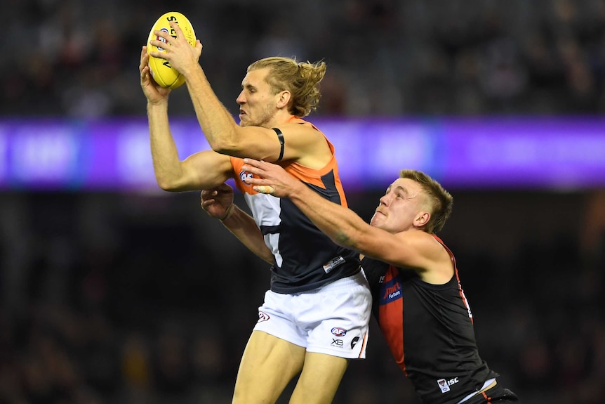 An AFL defender grasps the football above his head while an opponent tries to grab him from behind.