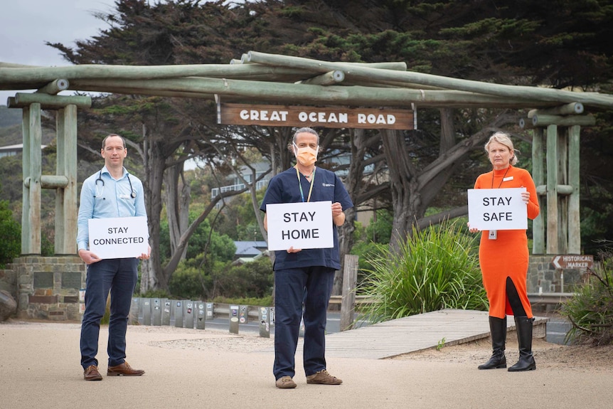 Two male and one female GP holding signs urging people to stay away stand outside in front of a sign saying  "Great Ocean Road"