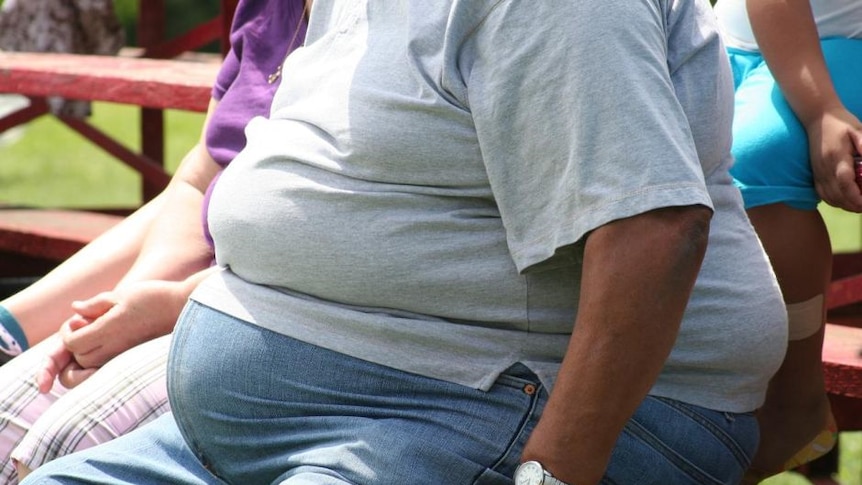 An obese person, wearing a grey t-shirt and jeans, sitting outside on a bench.