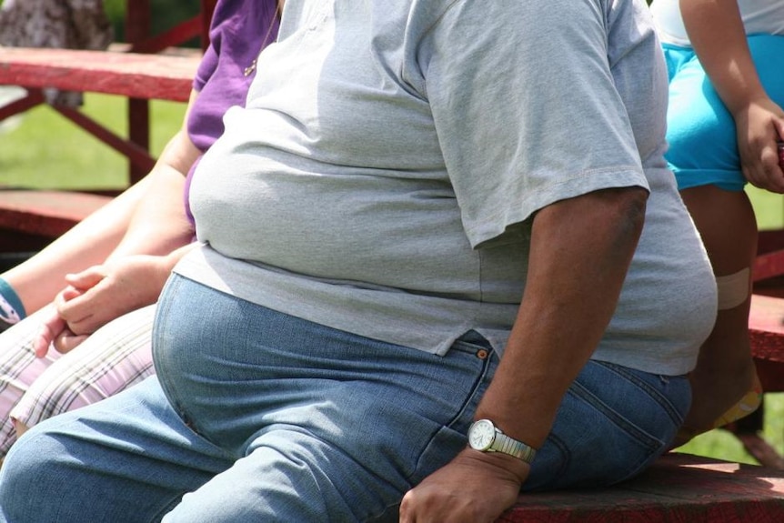 An obese person, wearing a grey t-shirt and jeans, sitting outside on a bench.