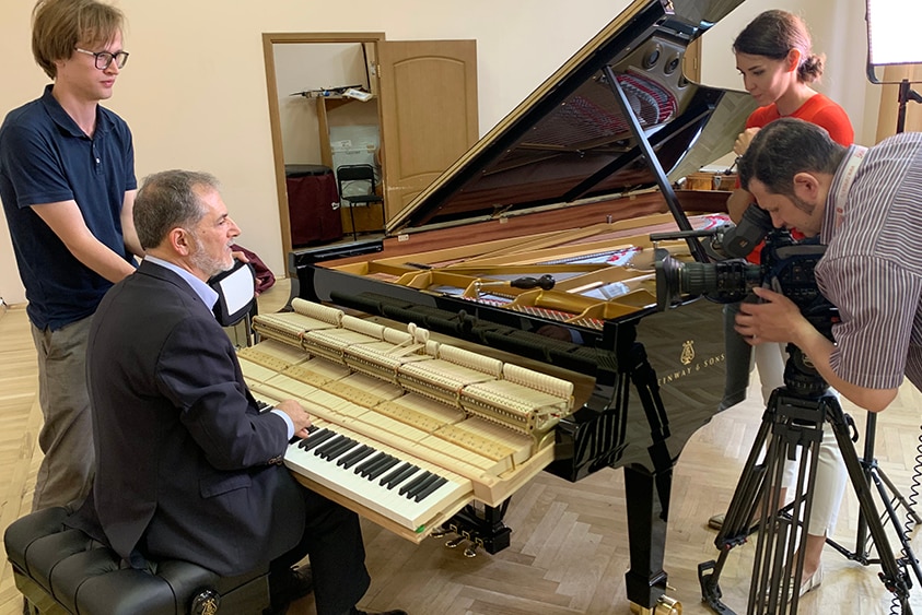 Piano technician Ara Vartoukian sits at a grand piano with the keyboard pulled out and film crew filming.