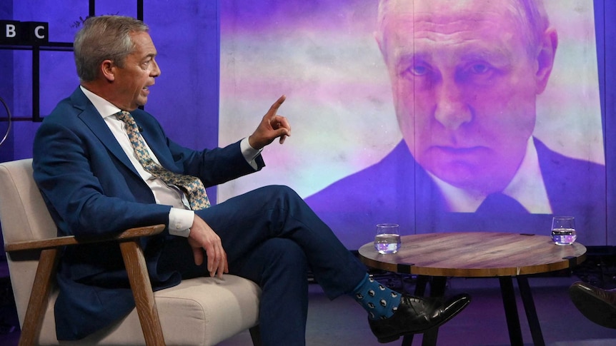 Nigel Farage sits in a brown armchair, pointing at a photo of Vladimir Putin.