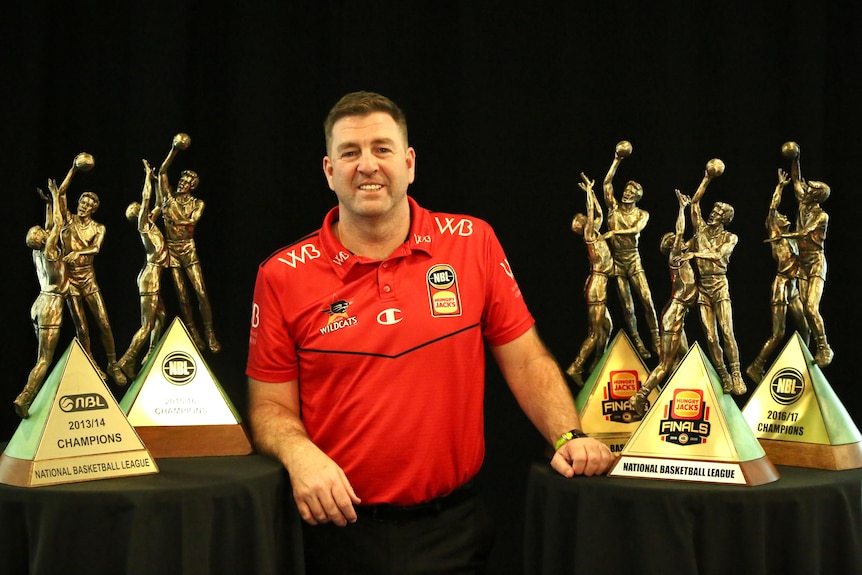 Trevor Gleeson wears a red shirt posing among five gold basketball trophies.
