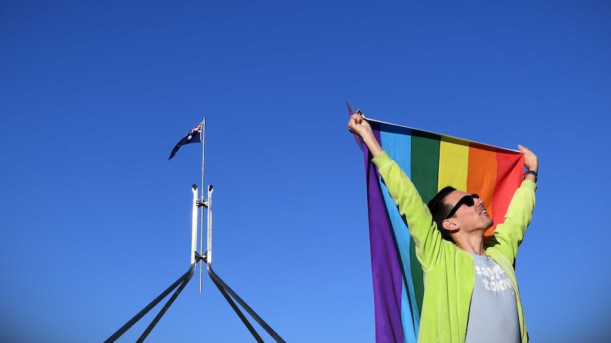 A man holds a rainbow flag above his head outside Parliament House.