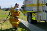 A young firefighter wearing bright orange fire safety uniform sprays water from a hose at a fire truck.