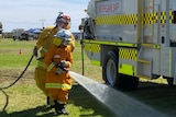 A young firefighter wearing bright orange fire safety uniform sprays water from a hose at a fire truck.