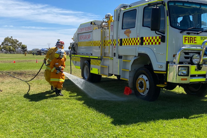 A young firefighter wearing bright orange fire safety uniform sprays water from a hose at a fire truck.
