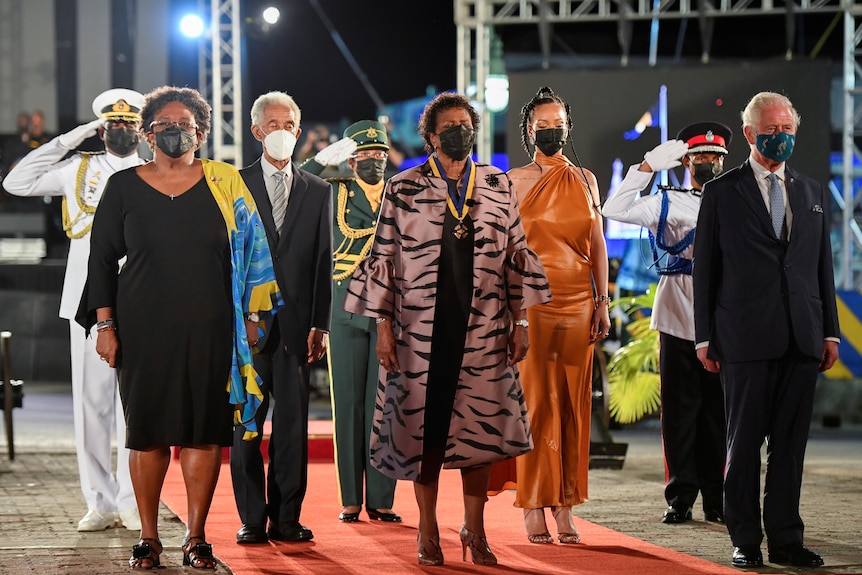 Sandra Mason, Mia Mottley and Prince Charles stand on red carpet. All are wearing formal attire and face masks
