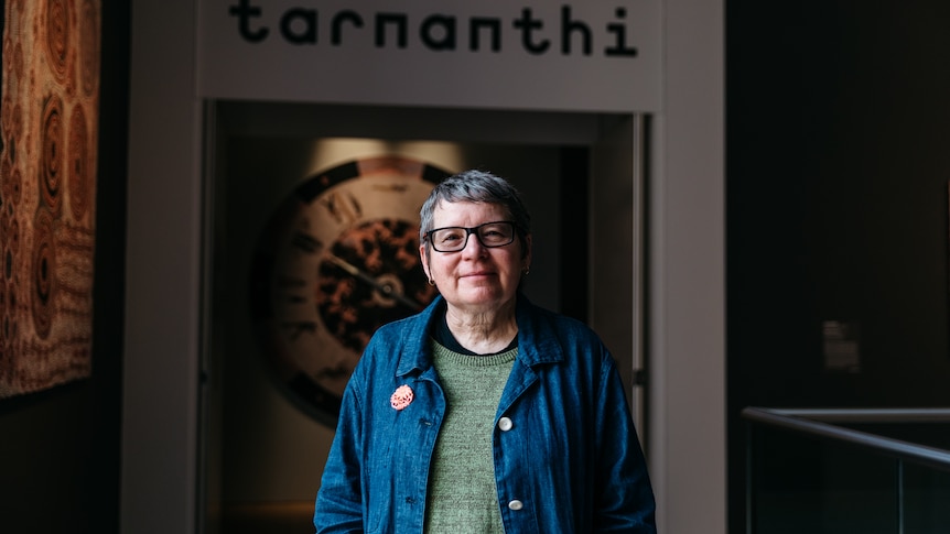 An Aboriginal woman with glasses and grey hair stands in front of a gallery with the words 'tarnanthi'