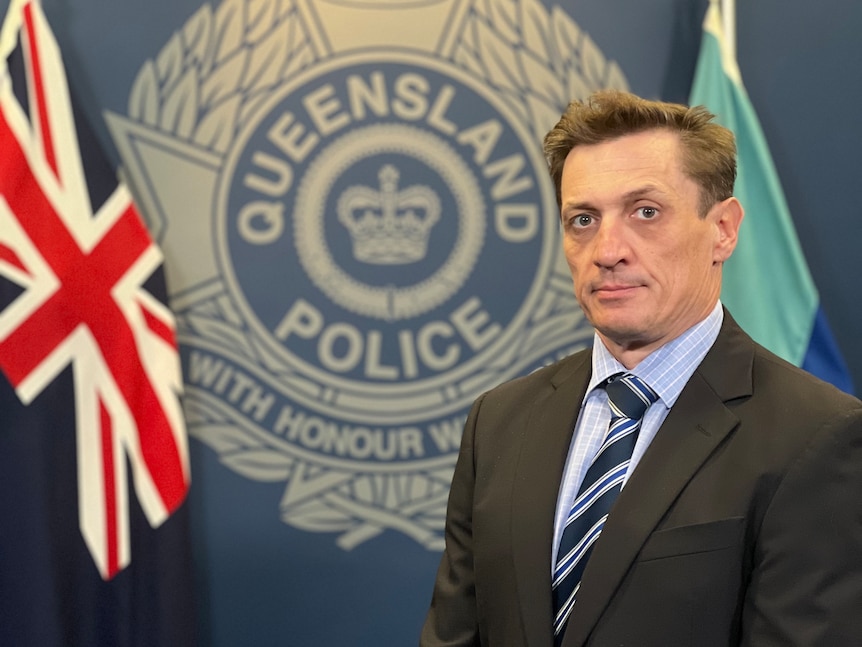 A man in a suit and tie with a blue shirt stands infront of the Queensland Police emblem and an Australian flag. 