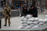 Two people in winter clothes prepare a barrier made of sandbags, as a person in military uniform stands nearby.