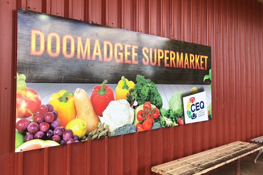 A sign, mounted on a corrugated tin wall, featuring photos of vegetables, reads 'Doomadgee supermarket'.