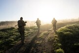 Three Ukrainian soldiers walk along a track as sunlight streams in from the horizon