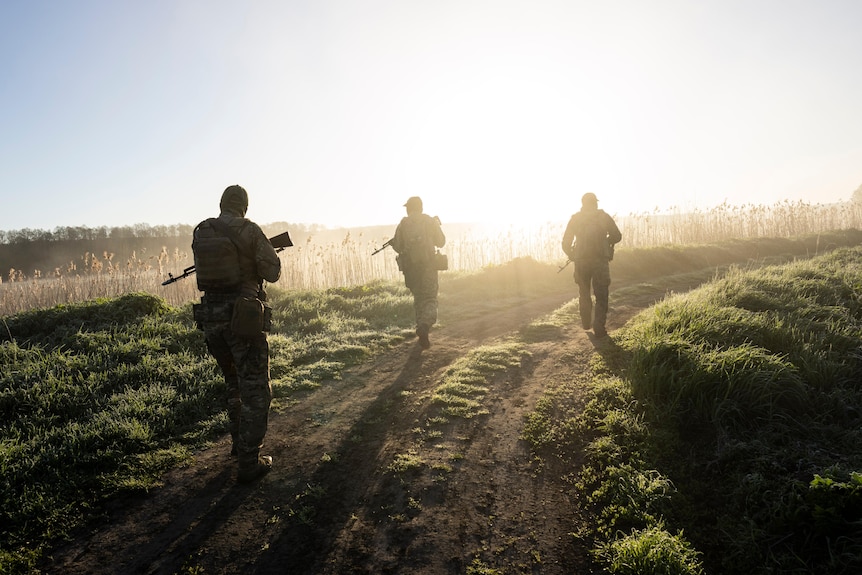 Three Ukrainian soldiers walk along a track as sunlight streams in from the horizon
