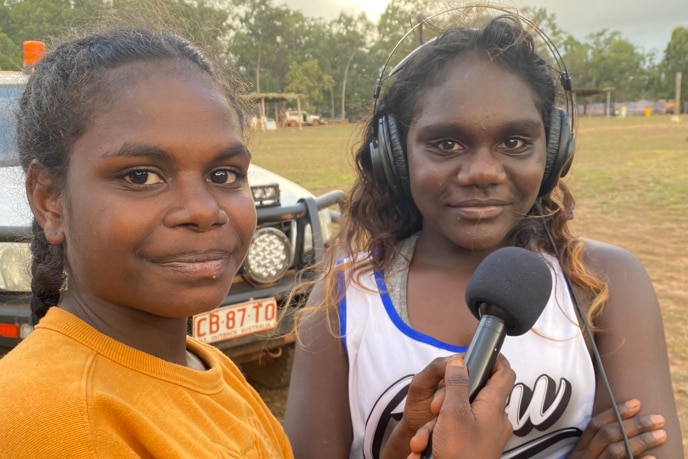 Two young girls standing on a field, one holding up a microphone as the other wears a set of headphones 