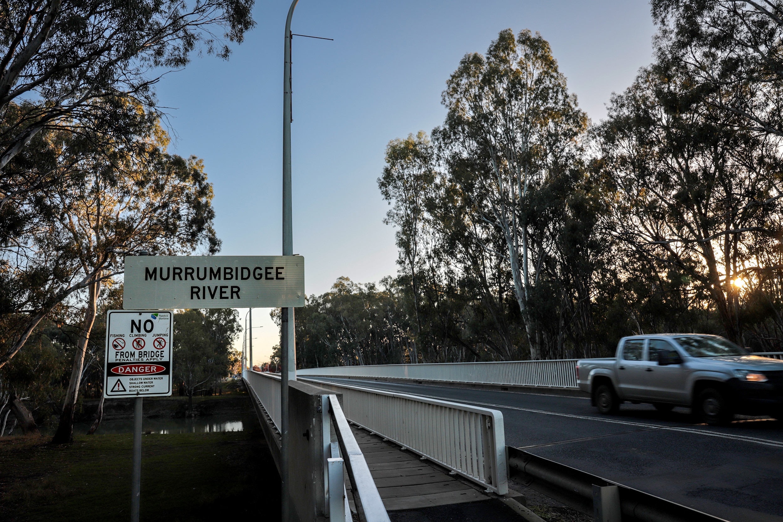 A sign next to a bridge reads 'MURRUMBIDGEE RIVER' a white utility vehicle passes on the opposite side of the road.