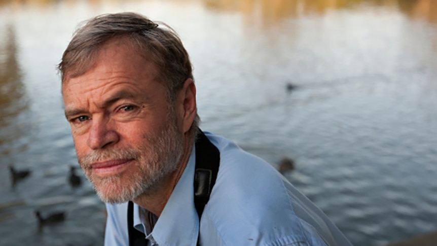 Richard Kingsford, dressed in a blue button-up shirt, kneels in front of a river.
