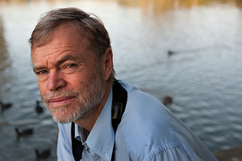 Richard Kingsford, dressed in a blue button-up shirt, kneels in front of a river.
