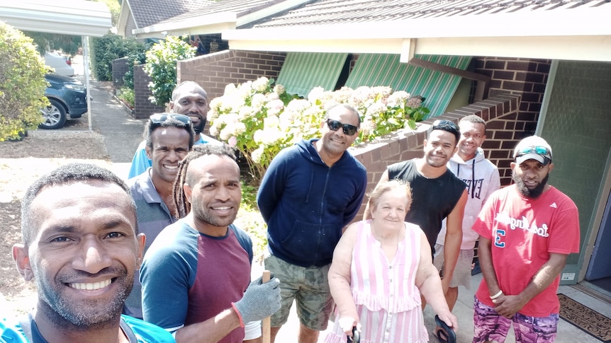 Group of eight men from Vanuatu smile at camera standing next to an elderly woman near brown brick building. 