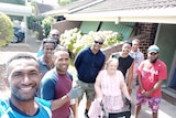 Group of eight men from Vanuatu smile at camera standing next to an elderly woman near brown brick building. 