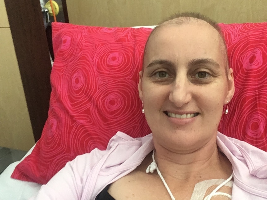 A woman with no hair smiles from a bed, with tubes attached to her.