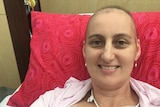 A woman with no hair smiles from a bed, with tubes attached to her.