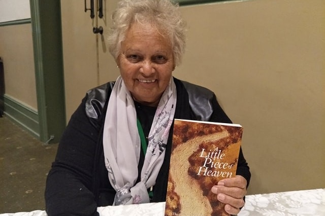 An older woman sitting down and holding a book to the camera.