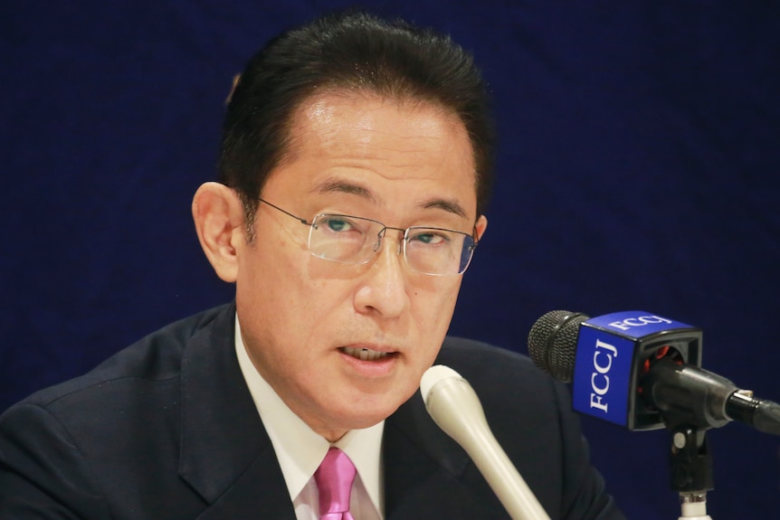 A man in glasses and a pink tie speaks into microphones.