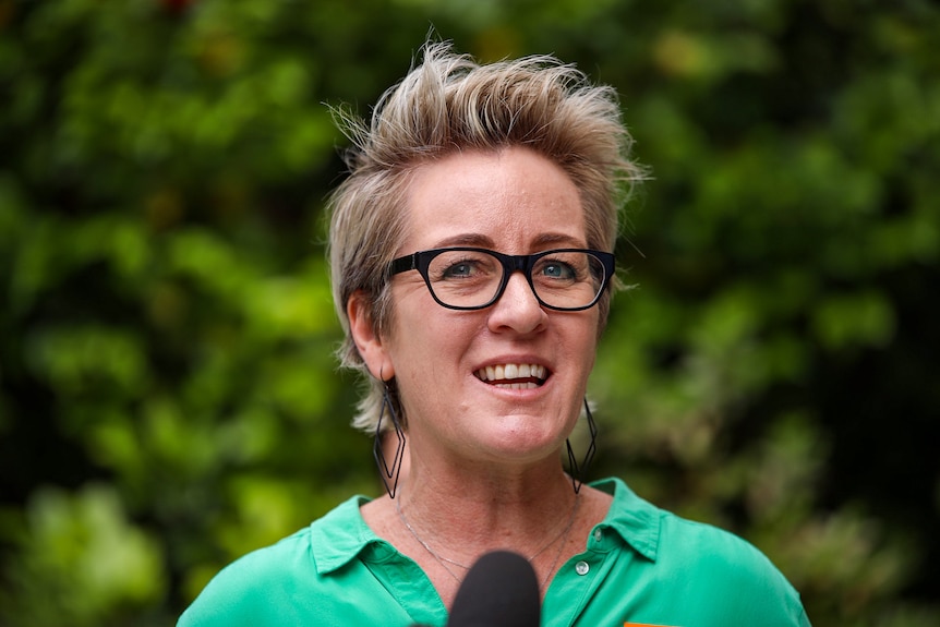 A woman with short blonde hair and a green shirt speaking at a press conference.
