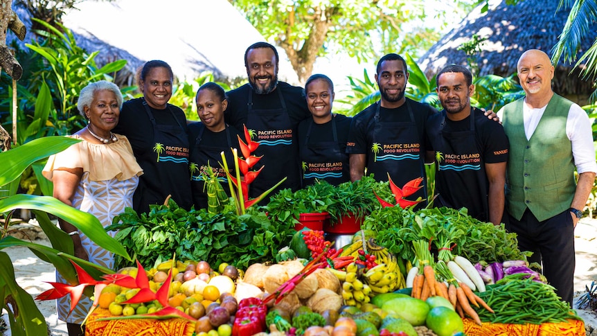 A group of Pacific Islanders stand together in black shirts in front of a table filled with vegetables