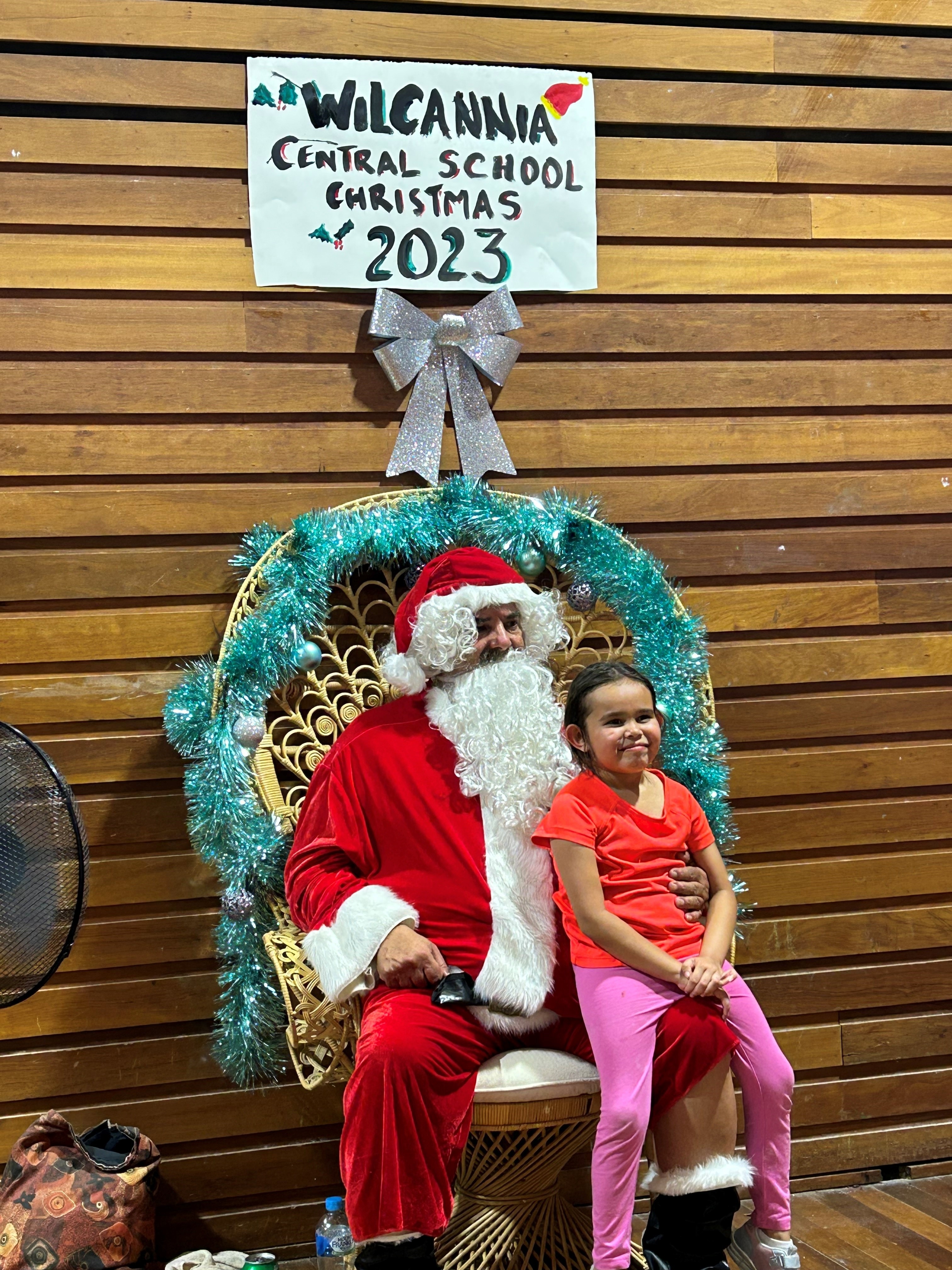 Santa sits in a big chair with a child on his knee who wears an orange shirt and pink pants.