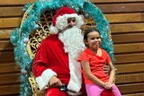 Santa sits in a big chair with a child on his knee who wears an orange shirt and pink pants.