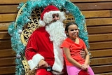 Santa sits in a big chair with a child on his knee who wears an orange shirt and pink pants.