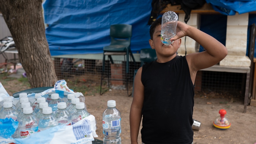 Young Indigenous boy stands next to a pallet of bottled water, drinking from one. 