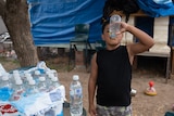 Young Indigenous boy stands next to a pallet of bottled water, drinking from one. 