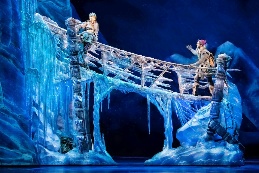 On a stage, a man and woman in winter clothes both stand on a frozen bridge bathed in blue light