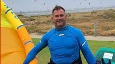 A man in a wetsuit holds an inflatable dinghy in one hand and a piece of outdoor equipment in the other.