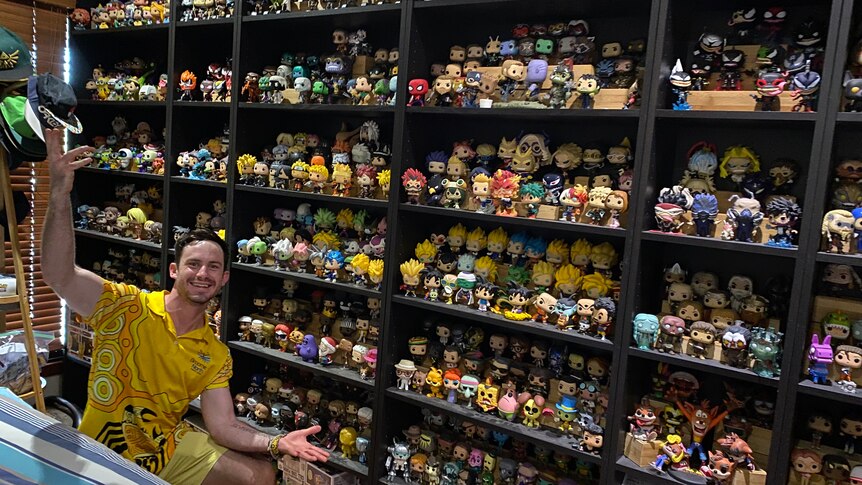 man on one knee with hands out to present his collection of pop vinyl figurines on shelves behind 