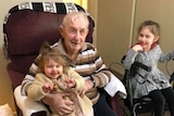 Alf Jordan sits in a red armchair, holding a beaming young girl on his knee while another girl sits in a walker smiling.