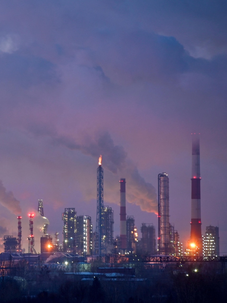 Clouds of gas and steam rise from chimneys and smokestacks of an oil refinery, while the sky is purple at dusk.