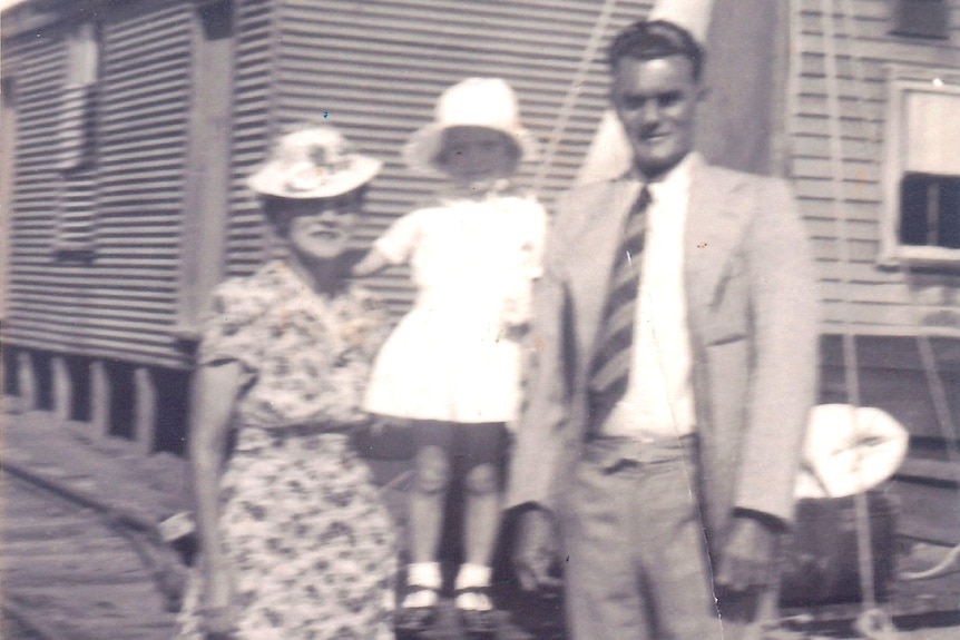 Black and white photo of man in suit, tie, woman wearing hat, printed dress, girl in white dress, hat in between, house behind.