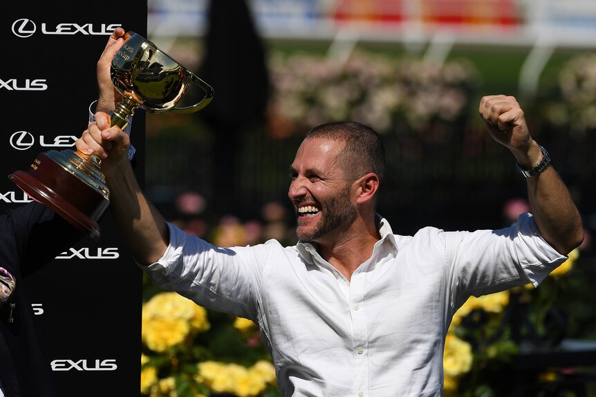 Two men hold up the Melbourne Cup happily in celebration.