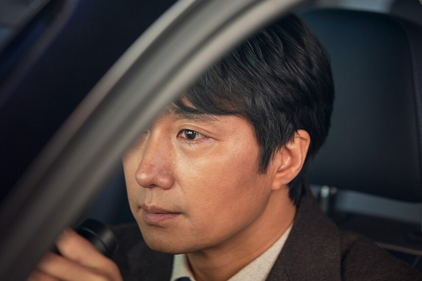 Close-up of East Asian man with black hair, wearing a black suit and tie, sitting in car with door ajar