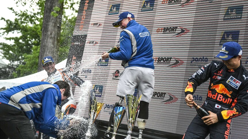 Billy Monger stands on the podium, spraying champagne at his teammates