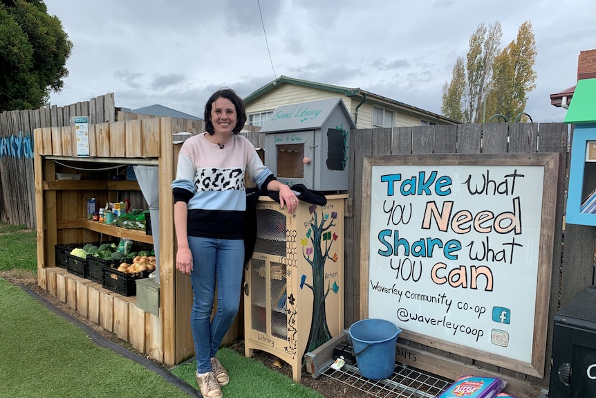 A woman stand next to a shelf of food and a sign saying, 'take what you need, share what you can'. 