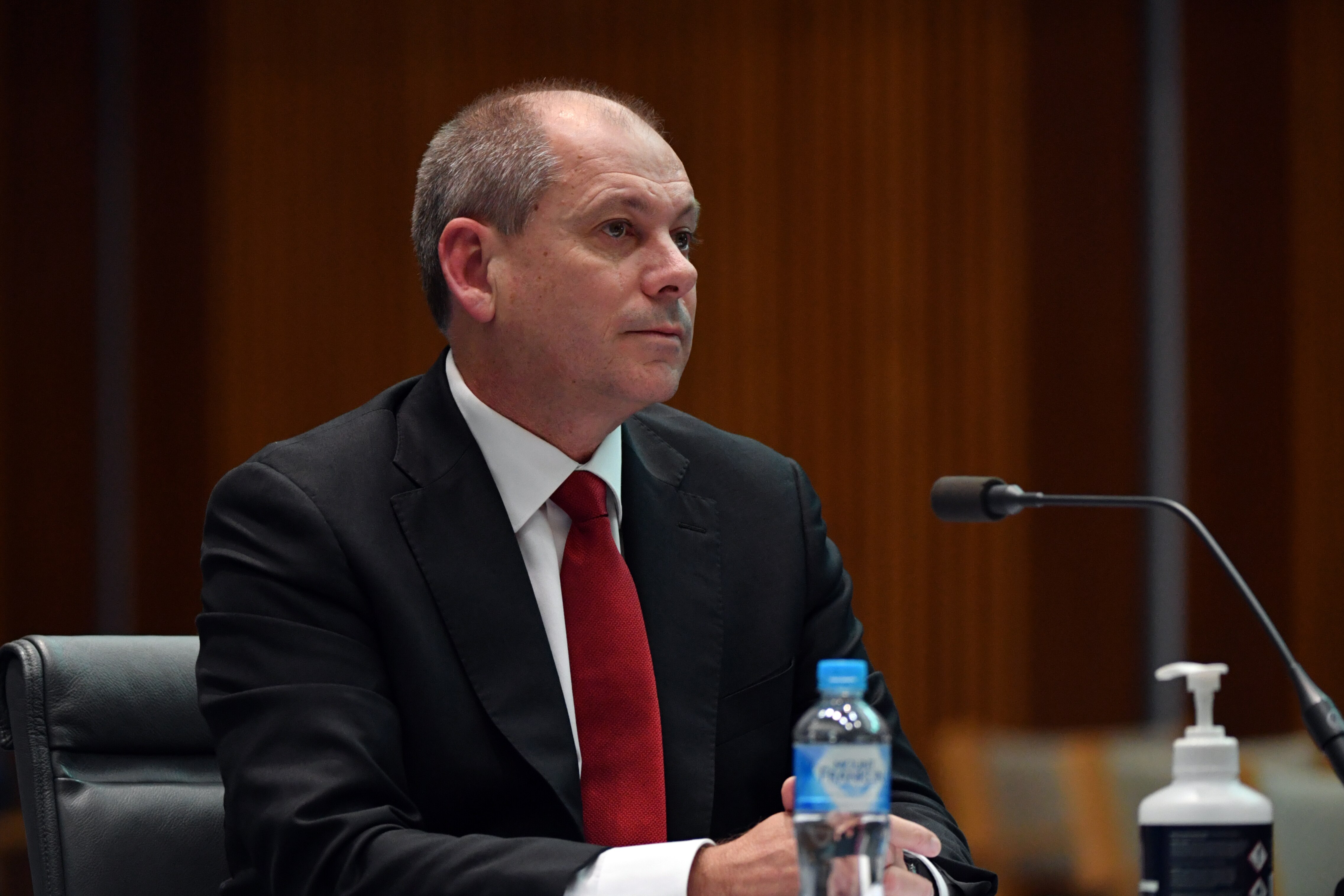A middle-aged white man in a suit with a red tie sits in a wood-panelled room behind a microphone.