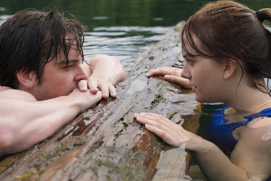 A teenage boy and girl are swimming. They gaze at each other over a log in the river.