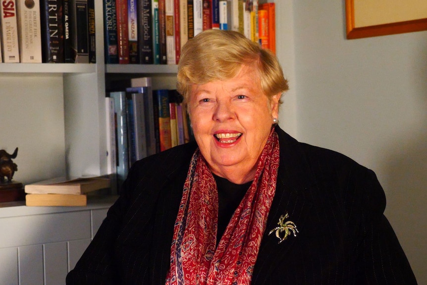 A lady in a black blazer and blonde hair looks at a camera