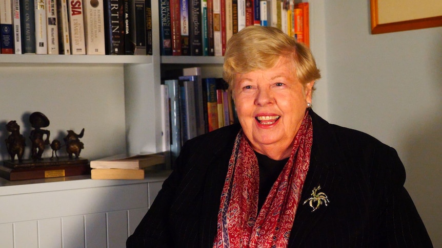 A lady in a black blazer and blonde hair looks at a camera